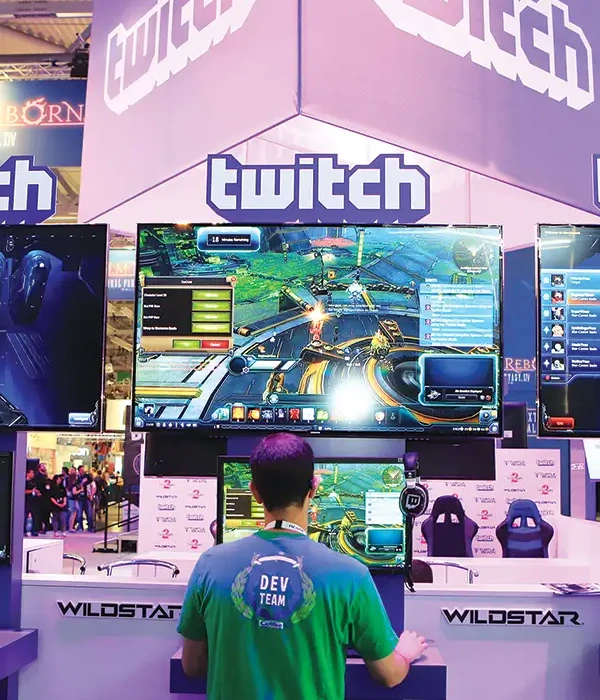 Developer demoing a new game under a giant Twitch banner at a convention, curious about buying Twitch followers to spark launch-day buzz.